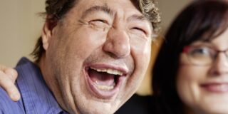 Zwei Menschen - ein Mann und eine Frau - sitzen vertraut und lachend am Tisch. Die Frau legt dem Mann eine Hand auf die Schulter.