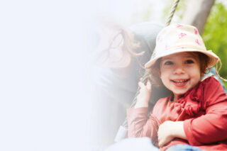 Ein Mädchen sitzt lachend in einer Nestschaukel, eine Erzieherin beugt sich fürsorglich zu ihr herunter.