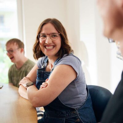 Junge Frau sitzt in einer Gruppe am Tisch, engagiert und lachend.