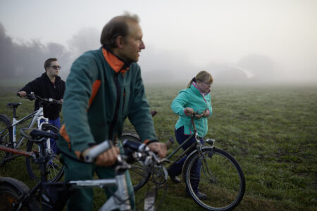 Drei Menschen - behindert und  nicht behindert -  schieben ihre Fahrräder in morgendlichem Dunst an einer Wiese.