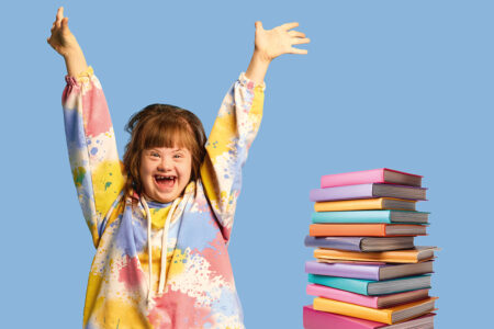 Ein Mädchen mit Down-Syndrom trägt einen bunten Jogginganzug und freut sich, neben ihr steht ein bunter Stapel Bücher.