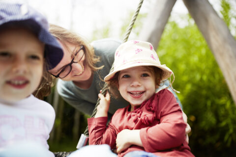 Kinder spielen lächelnd an einer Nestschaukel, eine Erzieherin beugt sich fürsorglich zu ihnen herunter.